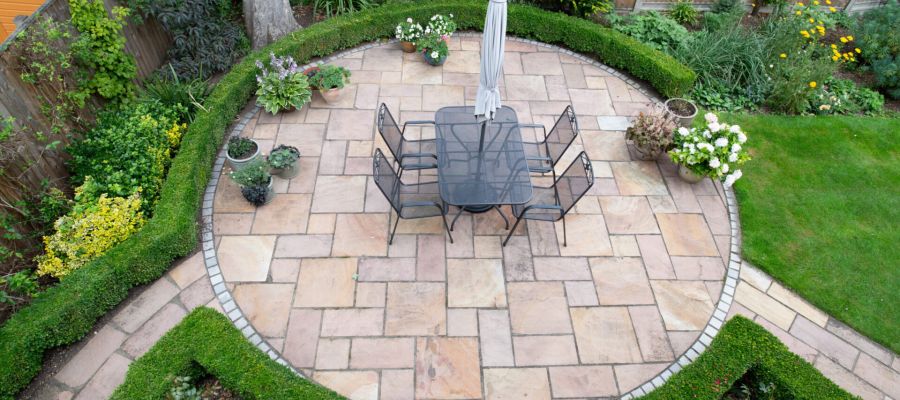 Circular patio garden with stone tiles and outdoor dining set, surrounded by lush greenery and flowers.