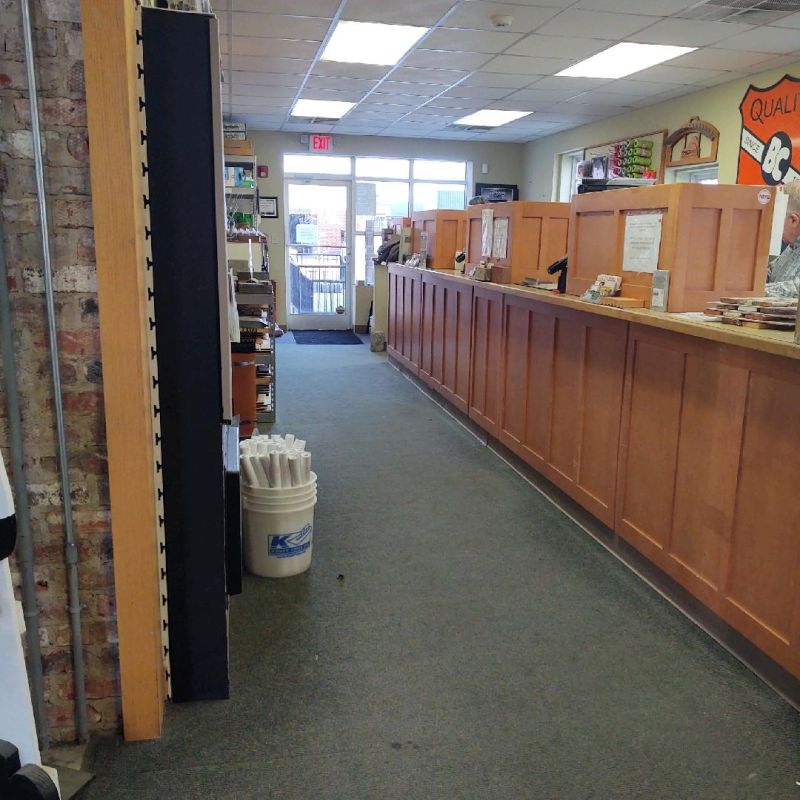 A store interior with a long wooden counter on the right, shelves on the left, and a glass door at the far end. The carpet is gray, and fluorescent lights illuminate the space. A Quality BC sign is above the counter.
