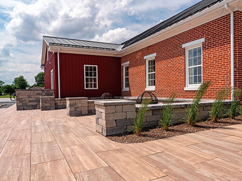 A brick building with white-framed windows, surrounded by a spacious patio with light brown tiles. Stone planters with grasses line the perimeter. The sky is partly cloudy, adding a bright backdrop.