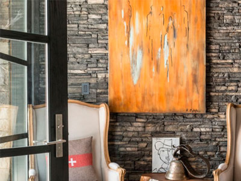 A cozy interior featuring a white armchair with a cushion, a small wooden table with a vintage bell, and an abstract orange painting on a textured stone wall. A glass door is partially open on the left.
