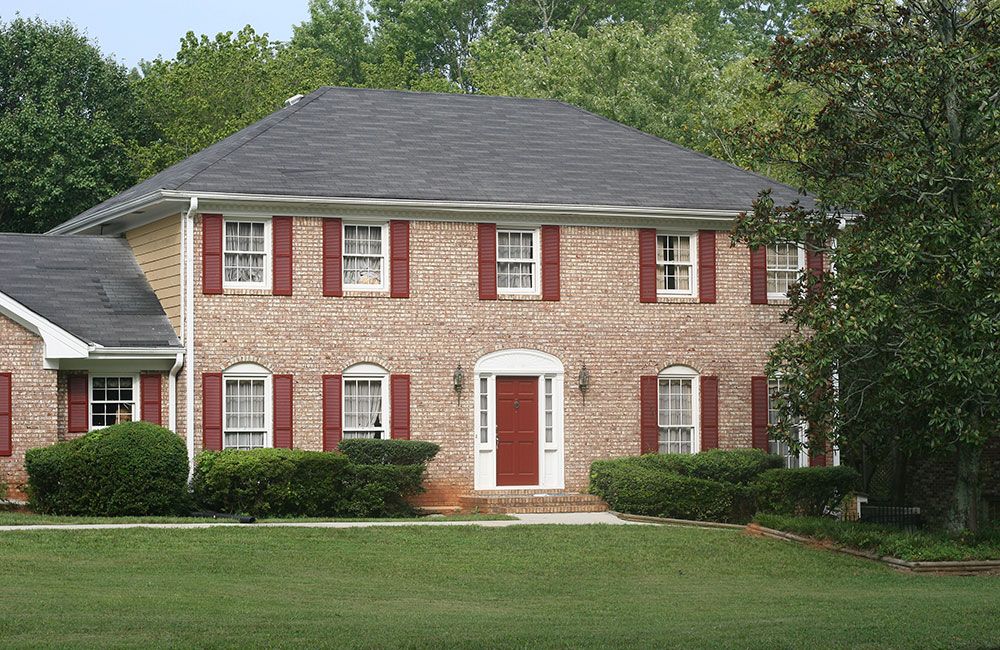 Two-story brick house with red shutters, a red door, and manicured shrubs, surrounded by green lawn and trees.