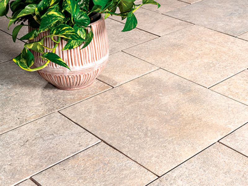 A potted plant with green leaves sits on a beige-tiled floor. The tiles have a smooth, slightly textured surface, arranged in a staggered pattern. The pot is ceramic with a ribbed texture.