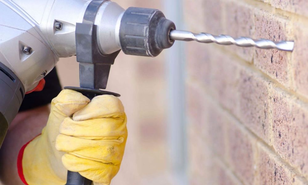 Person using a power drill with gloves to drill into a brick wall for construction.