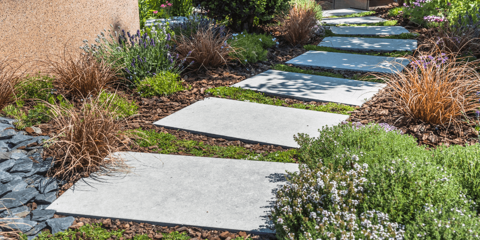 A garden path with large rectangular stone slabs leads through lush greenery, including shrubs and ornamental grasses. The path is bordered by small rocks and surrounded by various plants, creating a serene and inviting scene.
