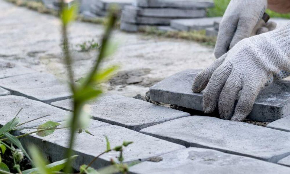 Gloved hands are laying gray paving stones in a row. Nearby, green grass and plants grow along the path, with more stones stacked in the background.