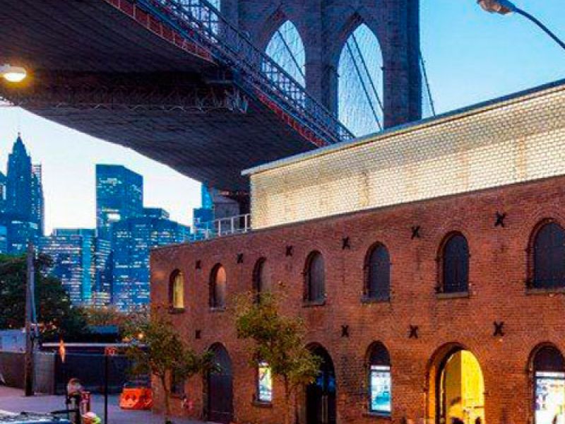 A street scene under the Brooklyn Bridge at dusk. Brick buildings line the road with arched windows illuminated by streetlights. Cars drive past as the city skyline is visible in the background against a dusky sky.