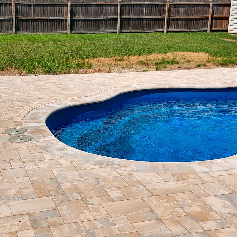 A backyard swimming pool with clear blue water is surrounded by a stone-tiled deck. In the background, there is a wooden fence and a patch of green grass.