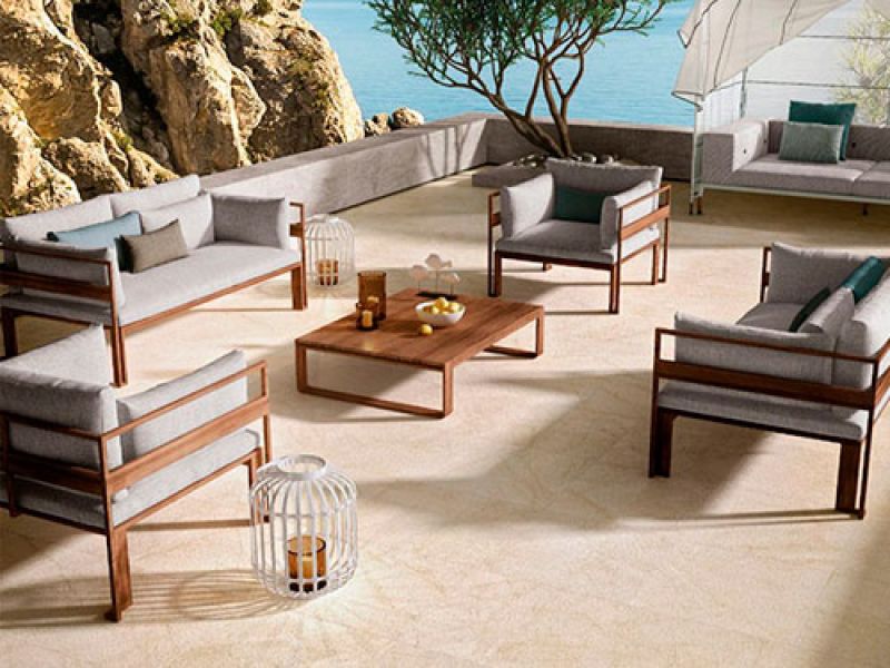 Outdoor patio with gray cushioned wooden furniture, including sofas and chairs, arranged around a rectangular table. A tree provides shade, and theres a view of the ocean and rocky cliffs in the background. Lanterns adorn the area.