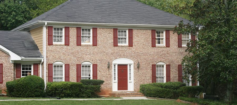 Two-story brick house with red shutters, a red door, and manicured shrubs, surrounded by green lawn and trees.