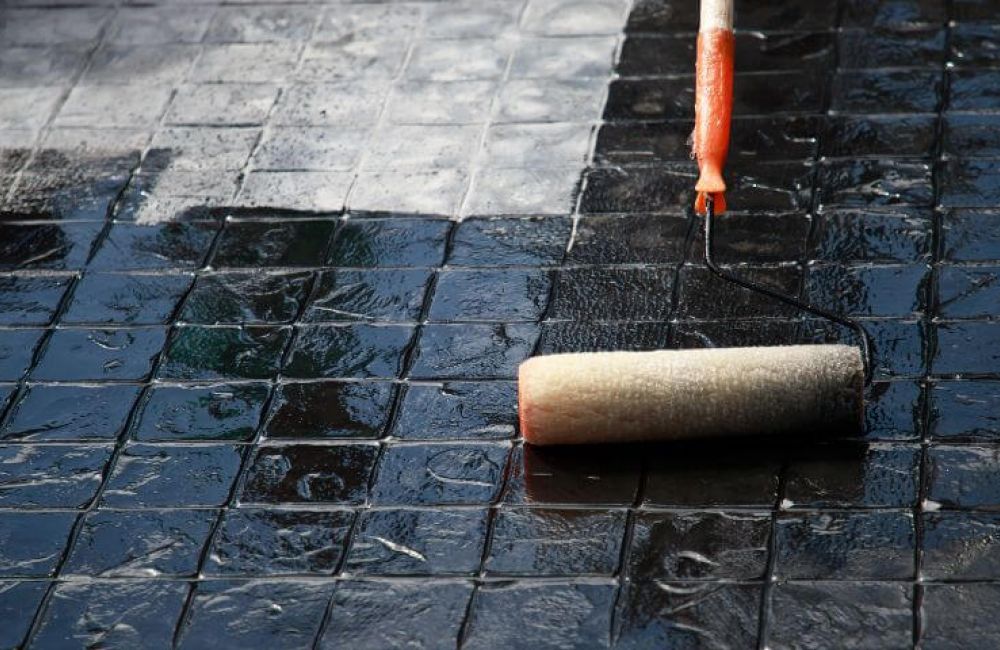 A paint roller applies a glossy black coating to textured tiles, reflecting a cloudy sky above.
