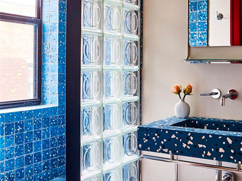 A bathroom with blue terrazzo tiles on a sink and wall, featuring a glass block divider. Theres a mirror above the sink and two orange flowers in a vase. Natural light streams in from a window on the left.