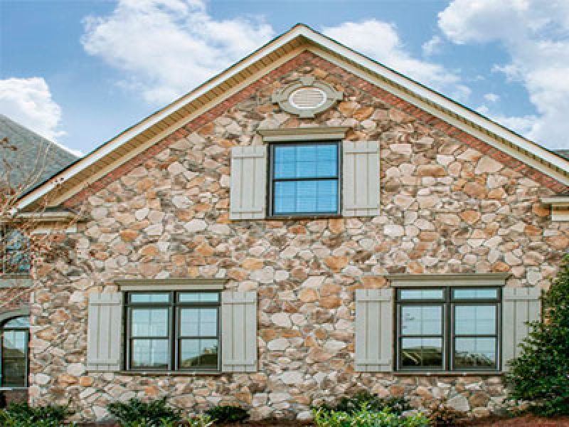 Front view of a stone house exterior with two windows. The windows have decorative shutters and the roof is shingled, with cloudy skies above. A small part of the roof and landscape are visible.