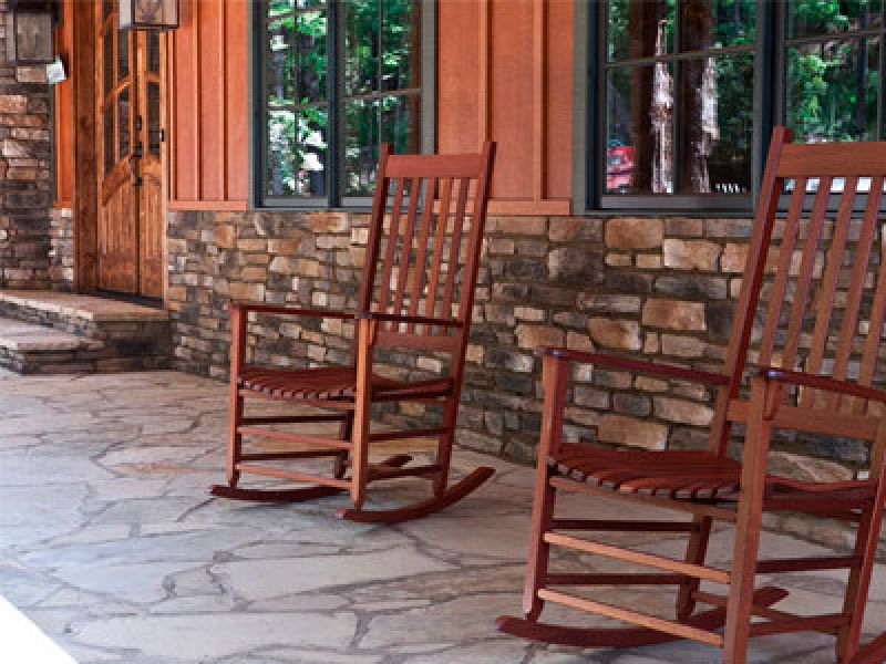Two wooden rocking chairs sit on a stone porch in front of a house with stone and wood exterior walls. Tall windows reflect lush green trees. A wooden door is visible on the left.
