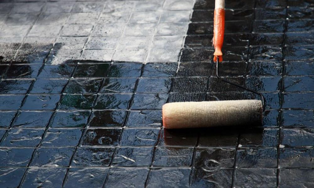 A paint roller applies a glossy black coating to textured tiles, reflecting a cloudy sky above.