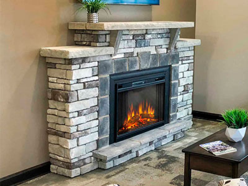 A stone fireplace with a modern electric fire feature is centered in a living room. The mantle holds a small plant and the hearth is surrounded by a cozy rug. A table with magazines and a potted plant is visible in the foreground.