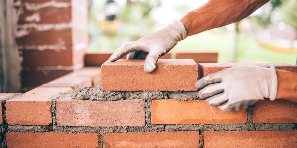 Hands wearing gloves lay a brick on mortar over an existing wall, demonstrating bricklaying in a construction setting.