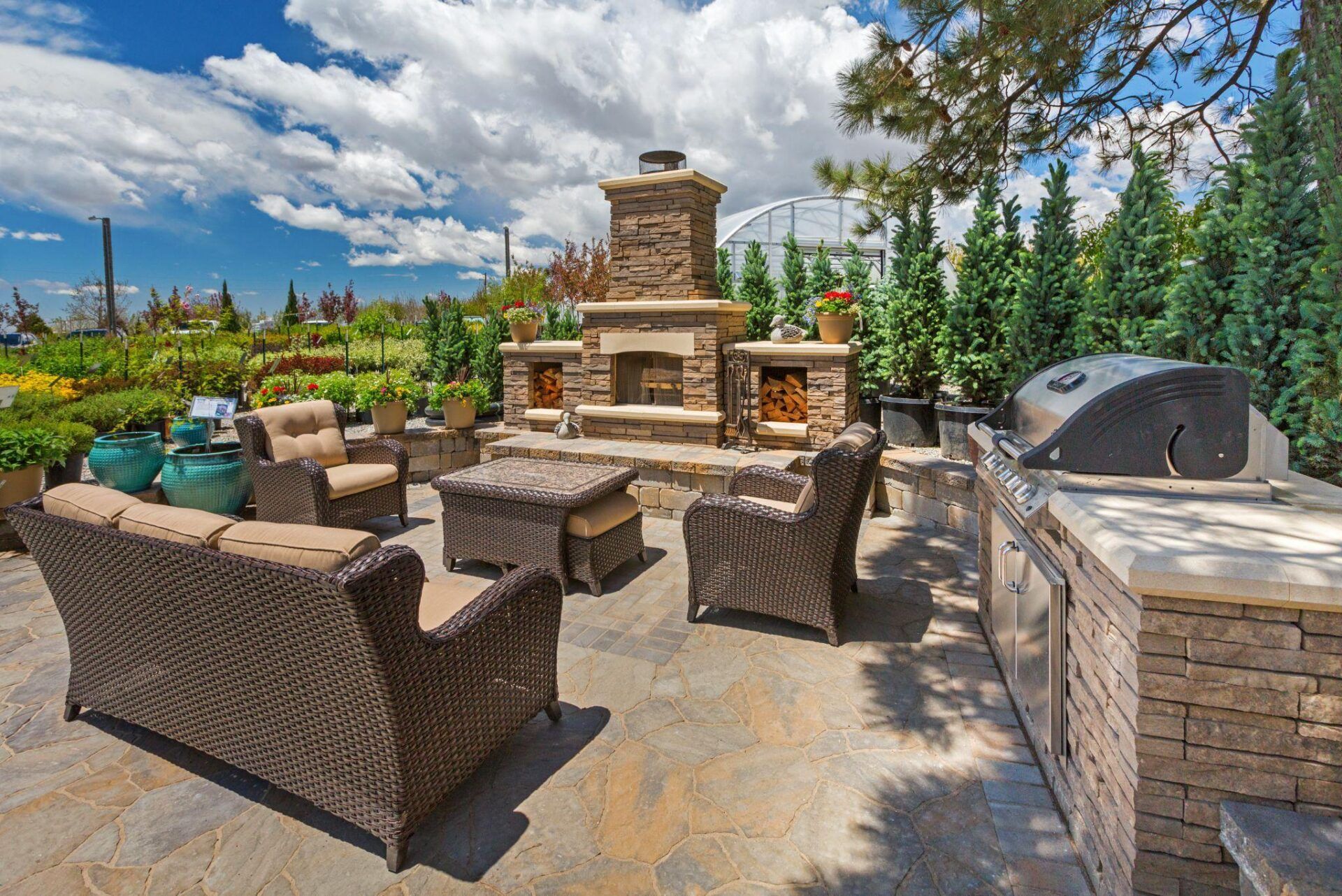 Outdoor patio with wicker furniture, stone fireplace, grill, and lush greenery under a blue sky.