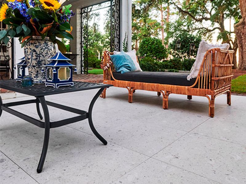 Patio scene with a wrought iron table holding a vase of colorful flowers and two small lanterns. In the background, theres a bamboo daybed with blue and white pillows, surrounded by lush greenery and a wrought iron gate.