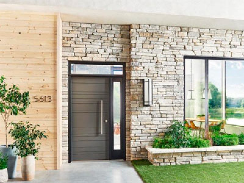 A modern entrance with a wooden wall and stone facade features a sleek black door. Large windows reveal an interior with a wooden table and chairs, surrounded by greenery and a view of a grassy outdoor area. House number 5613 is visible.