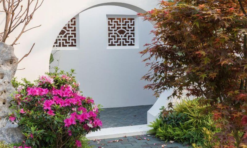 A garden with pink flowers and a stone, framed by a circular arch. Ornate windows sit in a white wall. Autumnal leaves add color, enhancing the serene, Asian-inspired setting.