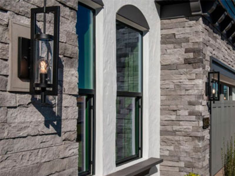 Exterior view of a house wall with modern stone cladding. Two black, rectangular outdoor wall lights are mounted beside arched and rectangular windows. The cladding features a mix of gray-toned stone.