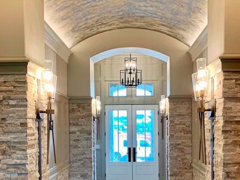 Elegant hallway with an arched ceiling and brick walls. Stylish light fixtures line the sides, and a modern chandelier hangs overhead. At the end, tall glass double doors lead outside, allowing natural light to brighten the space.