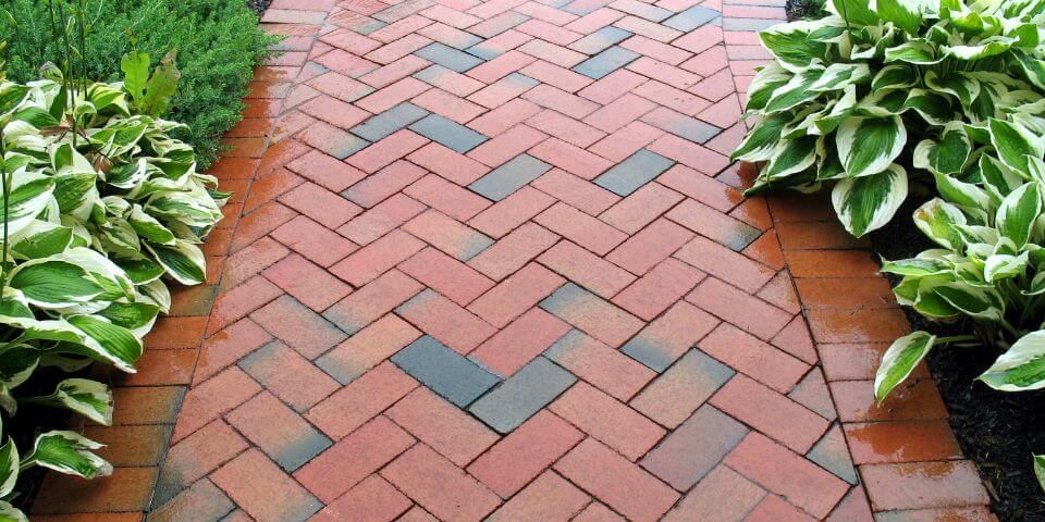 A patterned brick walkway lies between vibrant green foliage on either side, suggesting a well-maintained garden or park path.
