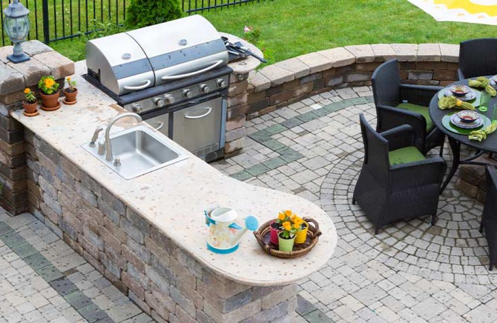 Outdoor kitchen with grill, sink, and patio dining area with round table and umbrella, surrounded by lush green grass.