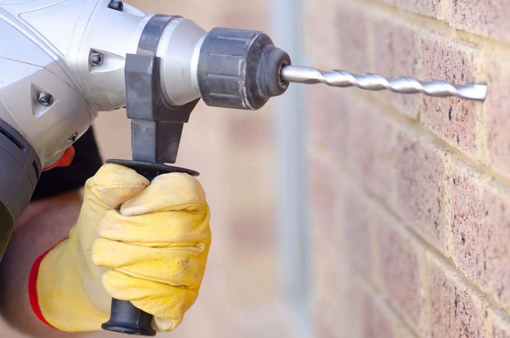Person using a power drill with gloves to drill into a brick wall for construction.
