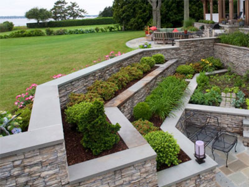 A terraced garden with stone walls features lush green shrubs and plants. There are outdoor seating areas with metal chairs and tables. In the background, a large grassy lawn leads to a view of water and trees.