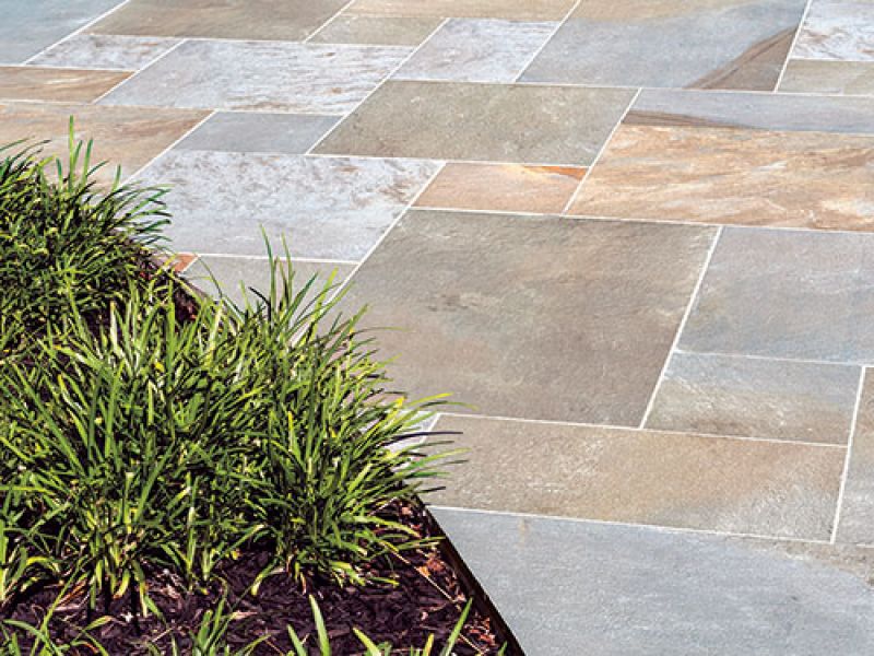 A neatly arranged stone patio with large rectangular tiles in various shades of grey and beige. A patch of green grass borders part of the patio, adding contrast to the clean, geometric lines of the stones.
