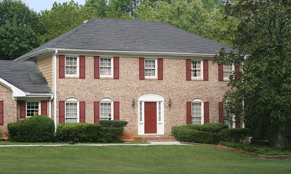 Two-story brick house with red shutters, a red door, and manicured shrubs, surrounded by green lawn and trees.
