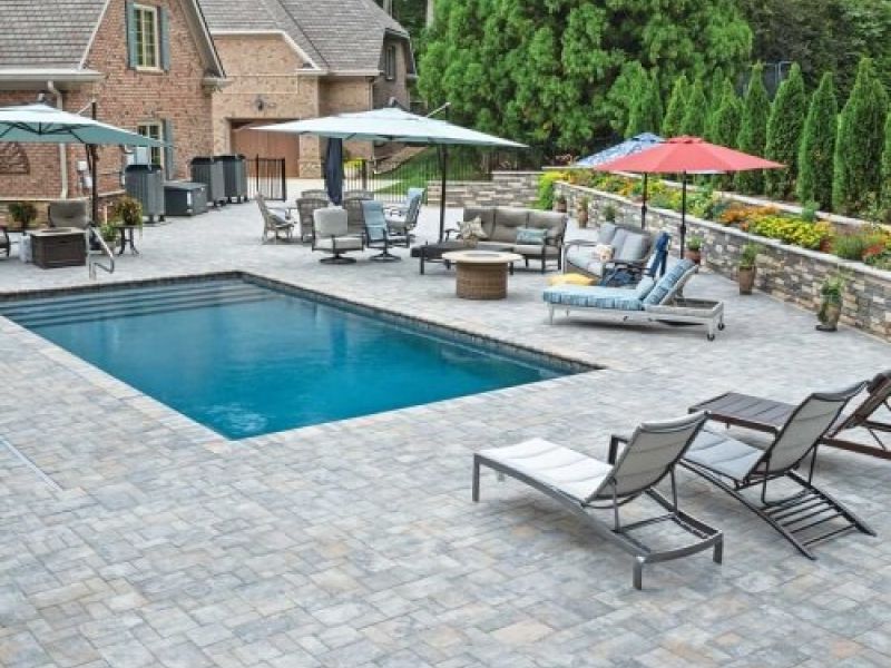 Luxury backyard with pool, loungers, umbrellas, and patio furniture surrounded by lush greenery and brick house.