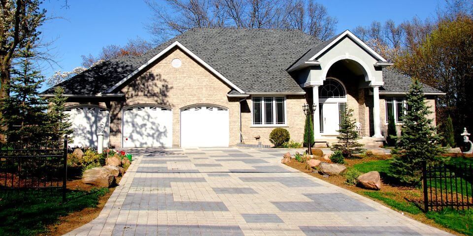 A single-story suburban house with a double garage sits under clear blue skies. A paved driveway leads up to it, flanked by manicured green lawns and ornamental bushes.