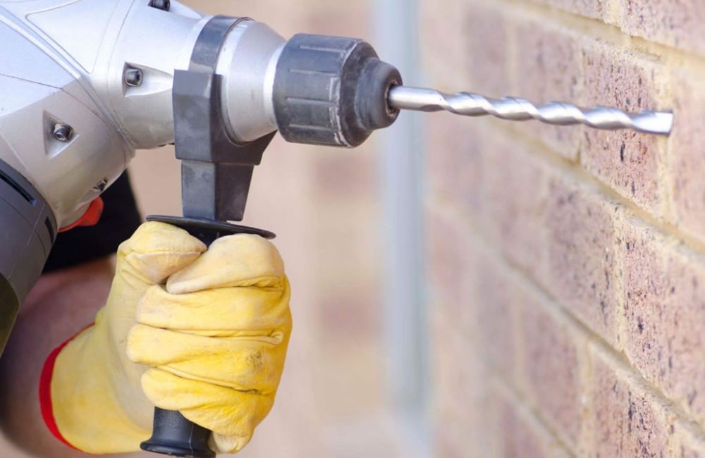 Person using a power drill with gloves to drill into a brick wall for construction.