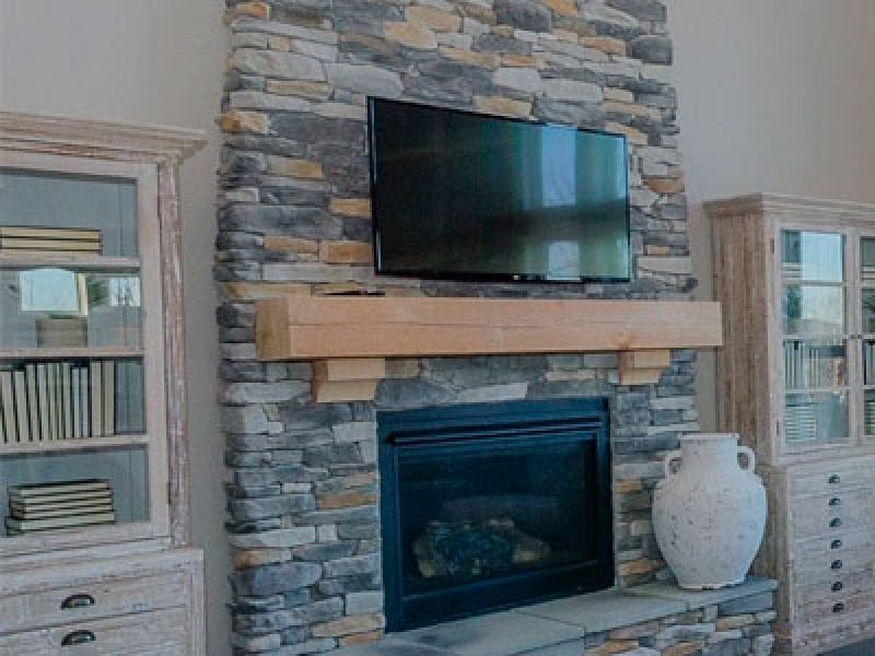 A rustic living room features a stone fireplace with a mounted flat-screen TV above. Wooden shelves flank the fireplace, filled with books. A large decorative vase sits on the hearth.