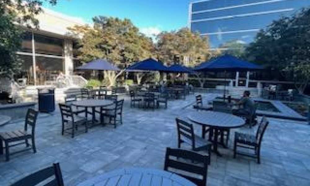 Outdoor patio with several round wooden tables and chairs under blue umbrellas. Few people are seated, surrounded by modern buildings and greenery. The sky is clear and sunny, creating a relaxing atmosphere.