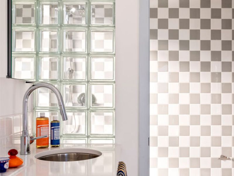 Modern bathroom with a sleek sink and chrome faucet on a white countertop. Glass block wall and checkered glass panel door partially visible. Fresh fruits and bottles adorn the counter.