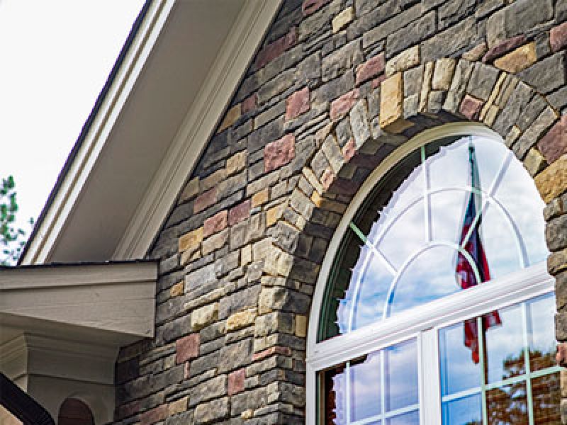 Close-up of a house exterior featuring a curved, multi-paned window with a reflective surface, framed by textured stonework in various earth tones. The reflection shows a blurred flag and trees in the glass.