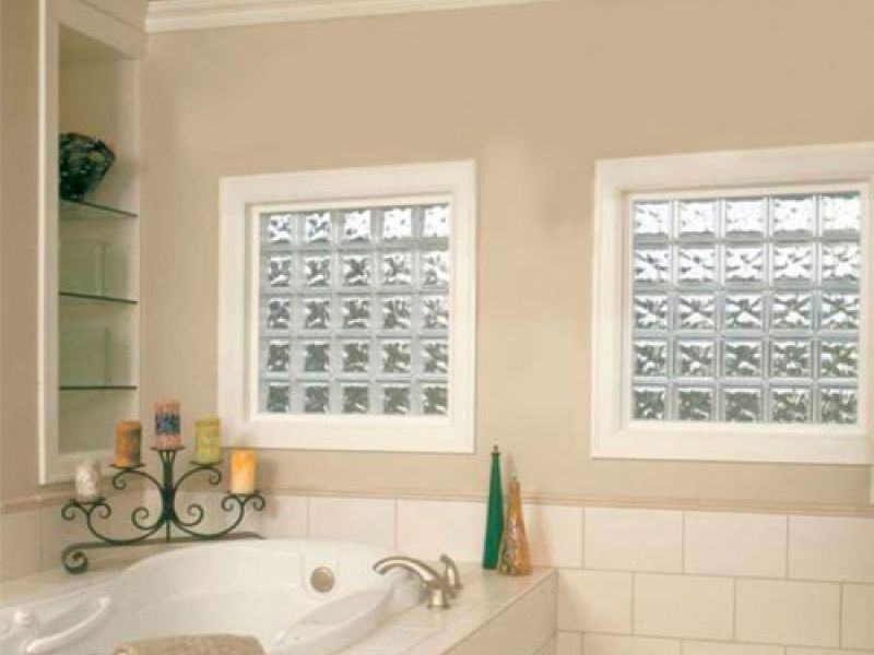 A bathroom with a large white bathtub surrounded by candles on a decorative stand. The walls are beige with two glass block windows. Towels are neatly placed on the tubs edge, and the floor is tiled in white.