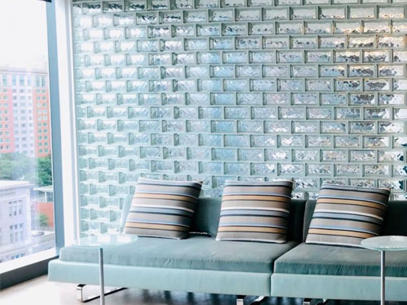 Modern lounge with a minimalist design featuring a light blue sofa with striped cushions, positioned against a wall of glass bricks. Two small round tables are nearby. Large windows offer a view of buildings and greenery.