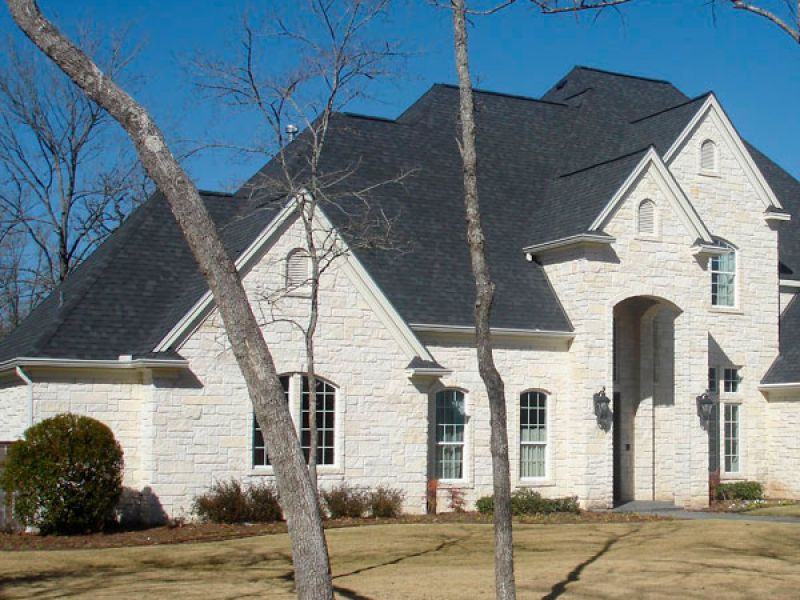 A two-story house with light stone exterior and dark gray roof on a clear day. Leafless trees and a dry grass lawn are in the foreground. The house has arched windows and a prominent entryway.