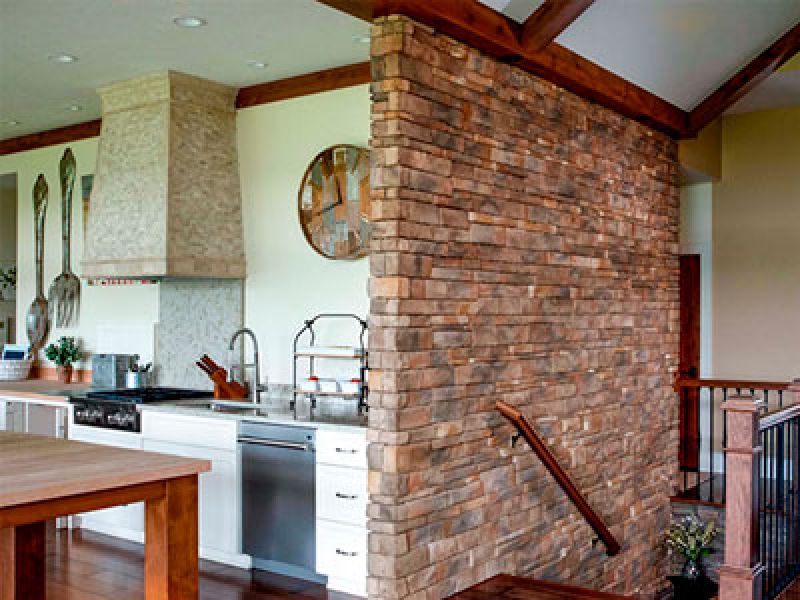 A modern kitchen featuring a large stone wall dividing a cooking area with a range and hood on the left from an open space on the right. The kitchen has a white countertop, a double sink, and utensils hanging on the wall. Wood beams accent the ceiling.