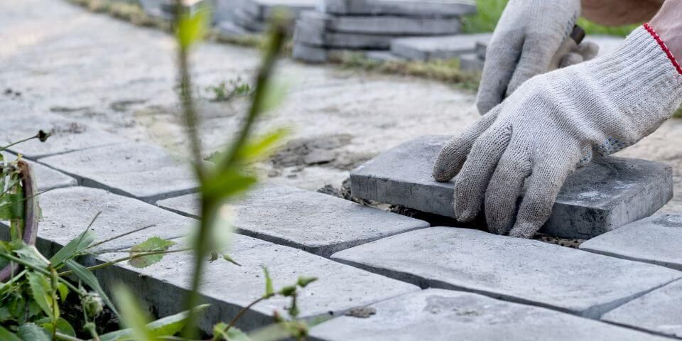 Gloved hands are laying gray paving stones in a row. Nearby, green grass and plants grow along the path, with more stones stacked in the background.