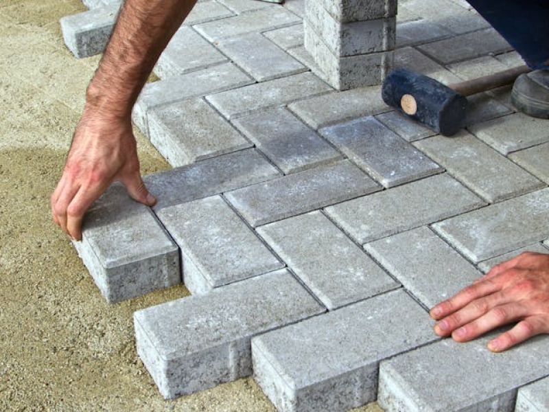Hands position gray bricks in a herringbone pattern on sandy ground, with a rubber mallet nearby, indicating ongoing pavement construction.