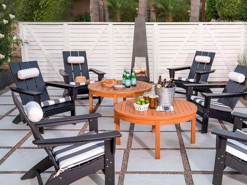 A patio scene with six black Adirondack chairs arranged around two round wooden tables topped with drinks, snacks, and a metal bucket of ice. A tall outdoor fireplace is in the background, with a white fence and greenery behind.