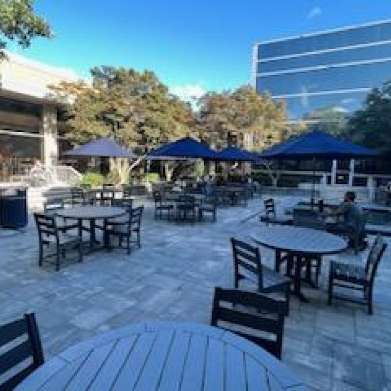 Outdoor patio with several round wooden tables and chairs under blue umbrellas. Few people are seated, surrounded by modern buildings and greenery. The sky is clear and sunny, creating a relaxing atmosphere.