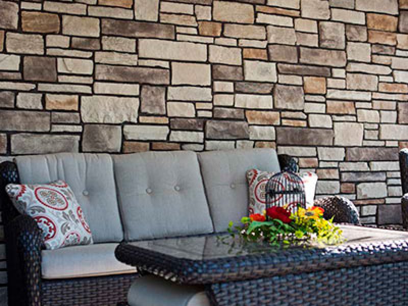 A cozy outdoor seating area features a wicker couch with gray cushions and patterned pillows in front of a stone wall. A glass-top table holds a birdcage and a colorful floral arrangement.