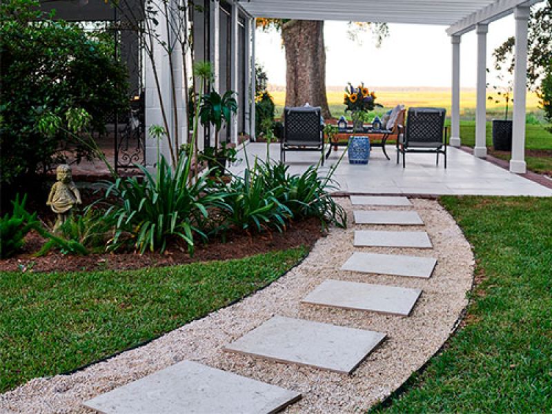 A garden patio with a pathway of square stone pavers leading to a sheltered seating area. The space is surrounded by lush greenery, with a small statue on the left. Two chairs and a table are on the patio, and a tree is visible in the background.