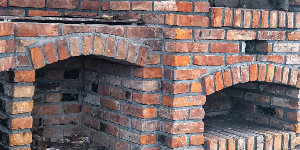 Brick oven structure with arched openings; located outdoors, the red and brown bricks form two separate oven chambers, set against a rustic environment with visible mortar joints.